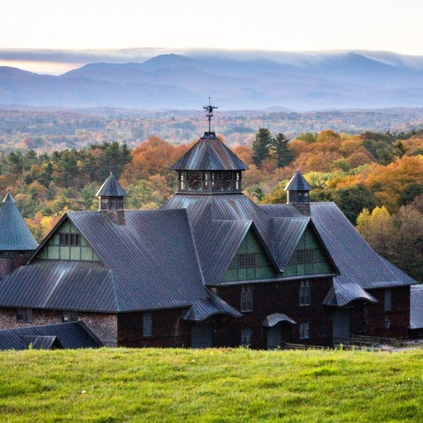 Historic farm building with distinctive metal roof and cupola set against a forested hillside with fall foliage